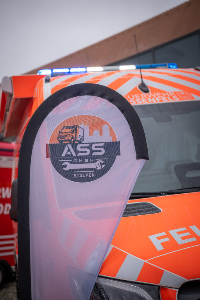 ASS-Logo auf einer Beachflag vor einem orangefarbenen Feuerwehr-Transporter